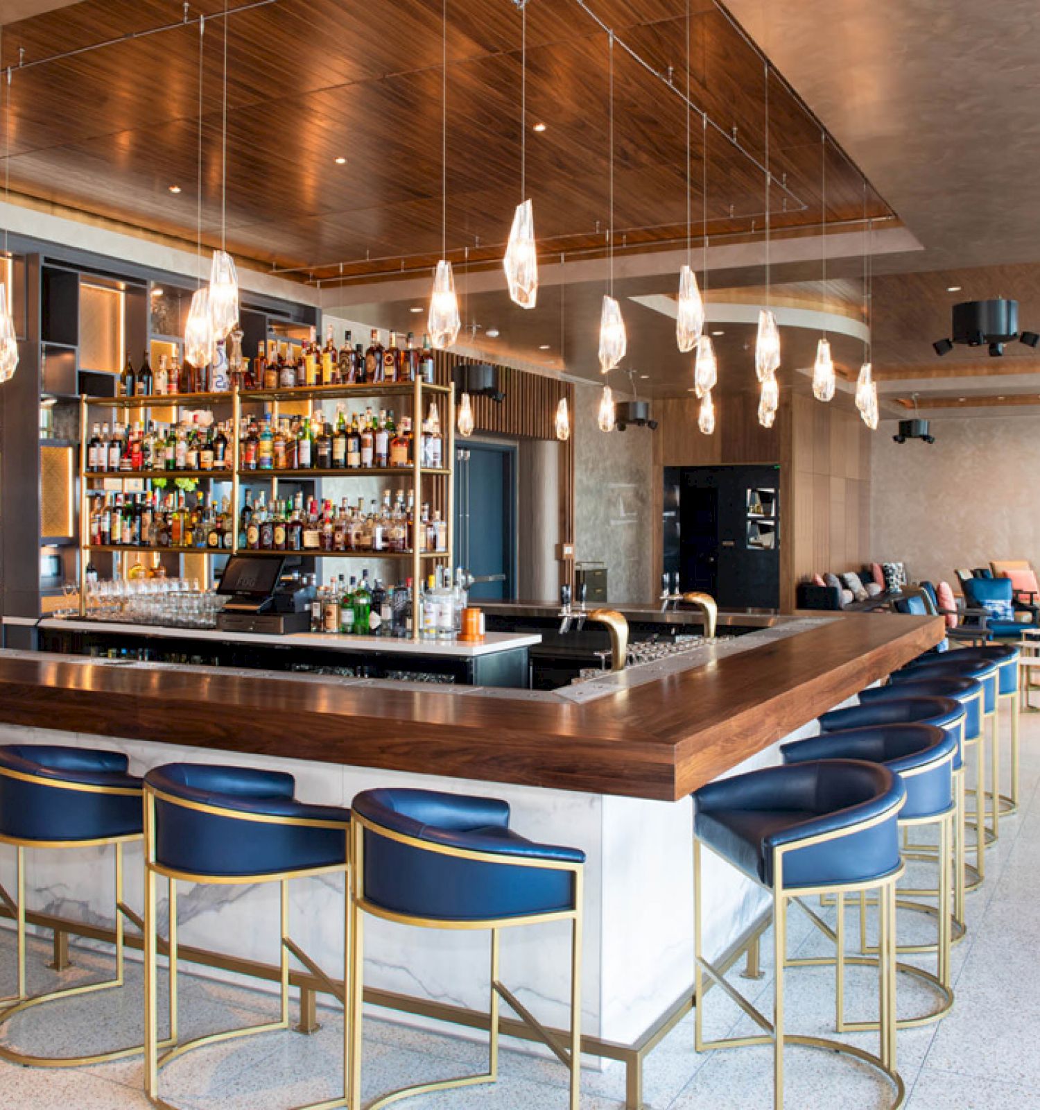 A modern bar with blue cushioned chairs, a wooden counter, hanging lights, and a well-stocked liquor shelf. Cozy seating is in the background.