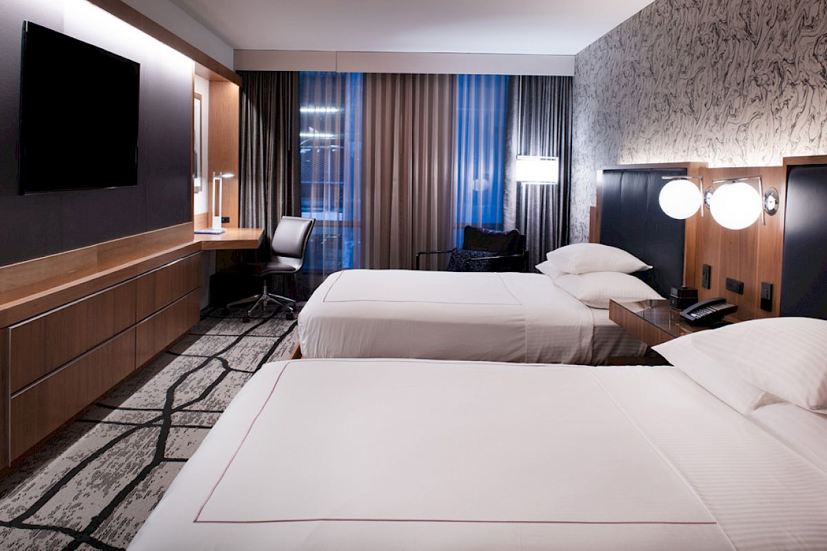 A modern hotel room with two beds, a flat-screen TV, desk and chairs, lamps, and stylish decor.