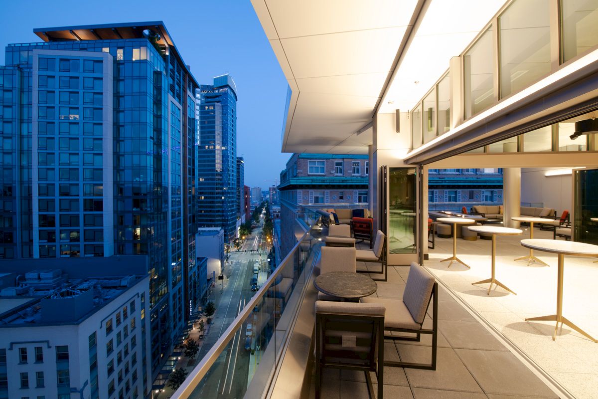 The image shows a modern rooftop terrace with seating, overlooking a city street and high-rise buildings at dusk.