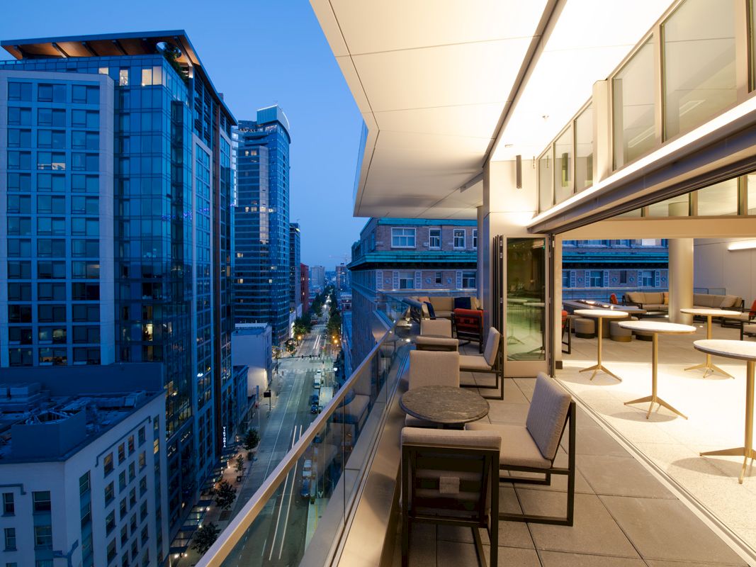 The image shows a modern rooftop terrace with seating, overlooking a city street and high-rise buildings at dusk.