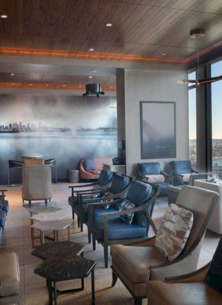 A modern lounge with stylish chairs and tables overlooks a waterfront view through large windows, featuring a skyline mural on the wall.