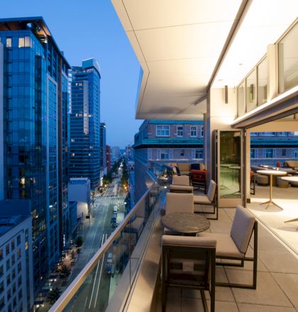 A modern urban balcony at dusk with tables and chairs overlooking a street and skyscrapers, offering a scenic view of the city skyline.