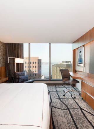 A modern hotel room with a large bed, television, desk, and window view of city buildings and water outside.