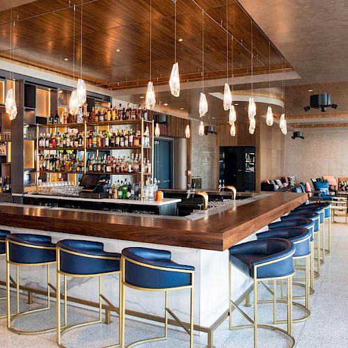 A modern bar with blue stools, hanging lights, and a stocked liquor shelf, in a spacious lounge with seating in the background.