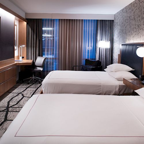 A modern hotel room with two beds, a desk, a TV, and stylish decor, featuring patterned wall and carpet, with large windows and curtains.