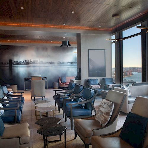 A modern lounge area with comfortable chairs, large windows offering a waterfront view, and a mural of a city skyline on the back wall.