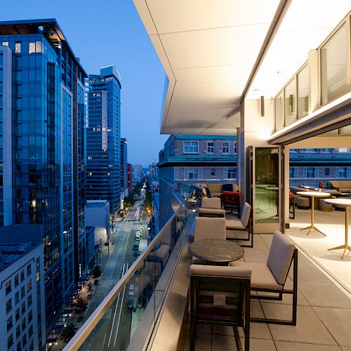 A modern city balcony with seating overlooks a street lined with tall buildings at dusk, creating an urban ambiance.