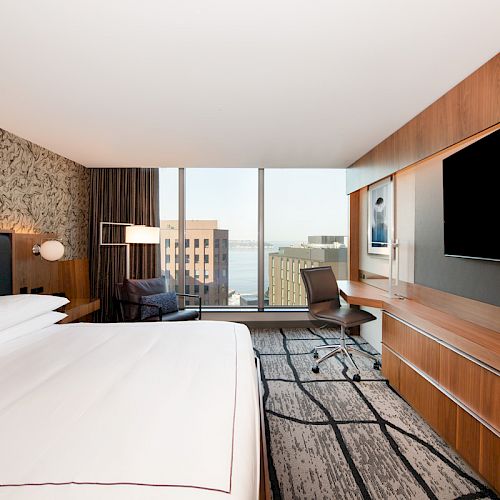 A modern hotel room with a large bed, TV, desk, and window view of buildings and water. Stylish decor and ample lighting complete the scene.