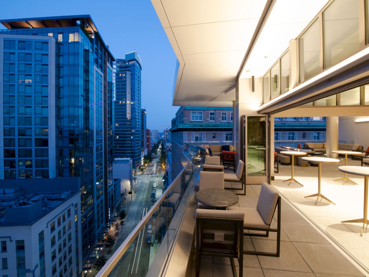 A modern balcony overlooking a cityscape at dusk, featuring tables and chairs, with tall buildings and street views.