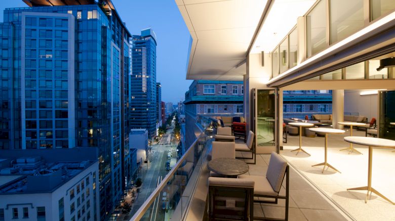 A modern balcony overlooking a cityscape at dusk, featuring tables and chairs, with tall buildings and street views.
