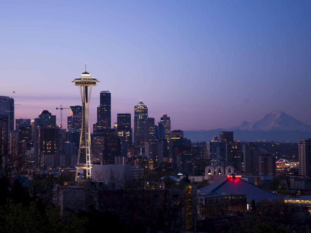 Seattle skyline at dusk with the Space Needle and Mount Rainier in the background, lit city lights against a twilight sky.