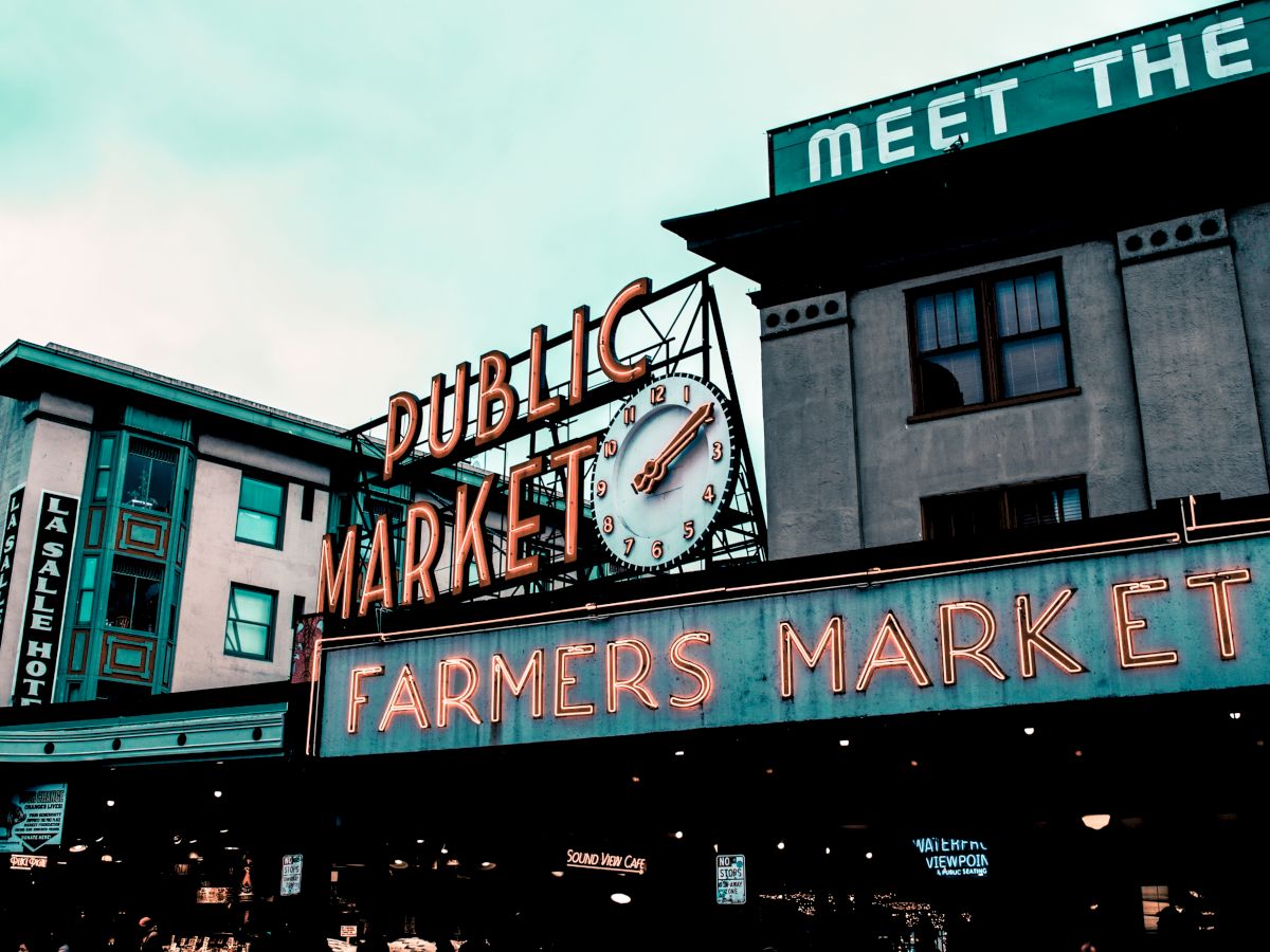 The image shows the iconic neon sign of Pike Place Market with a clock, featuring "Public Market" and "Farmers Market" in bold letters.