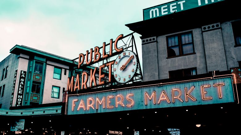 The image shows the iconic neon sign of Pike Place Market with a clock, featuring "Public Market" and "Farmers Market" in bold letters.
