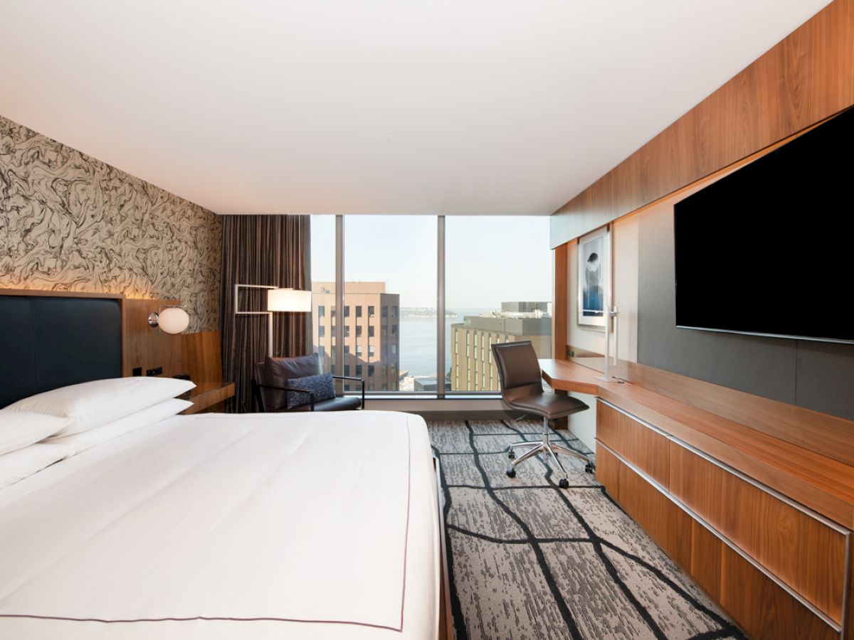 A modern hotel room with a large bed, desk, chair, and a flat-screen TV; wide window with a cityscape view in the background.