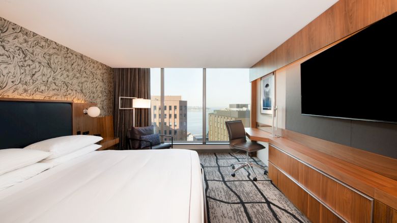 A modern hotel room with a large bed, desk, chair, and a flat-screen TV; wide window with a cityscape view in the background.