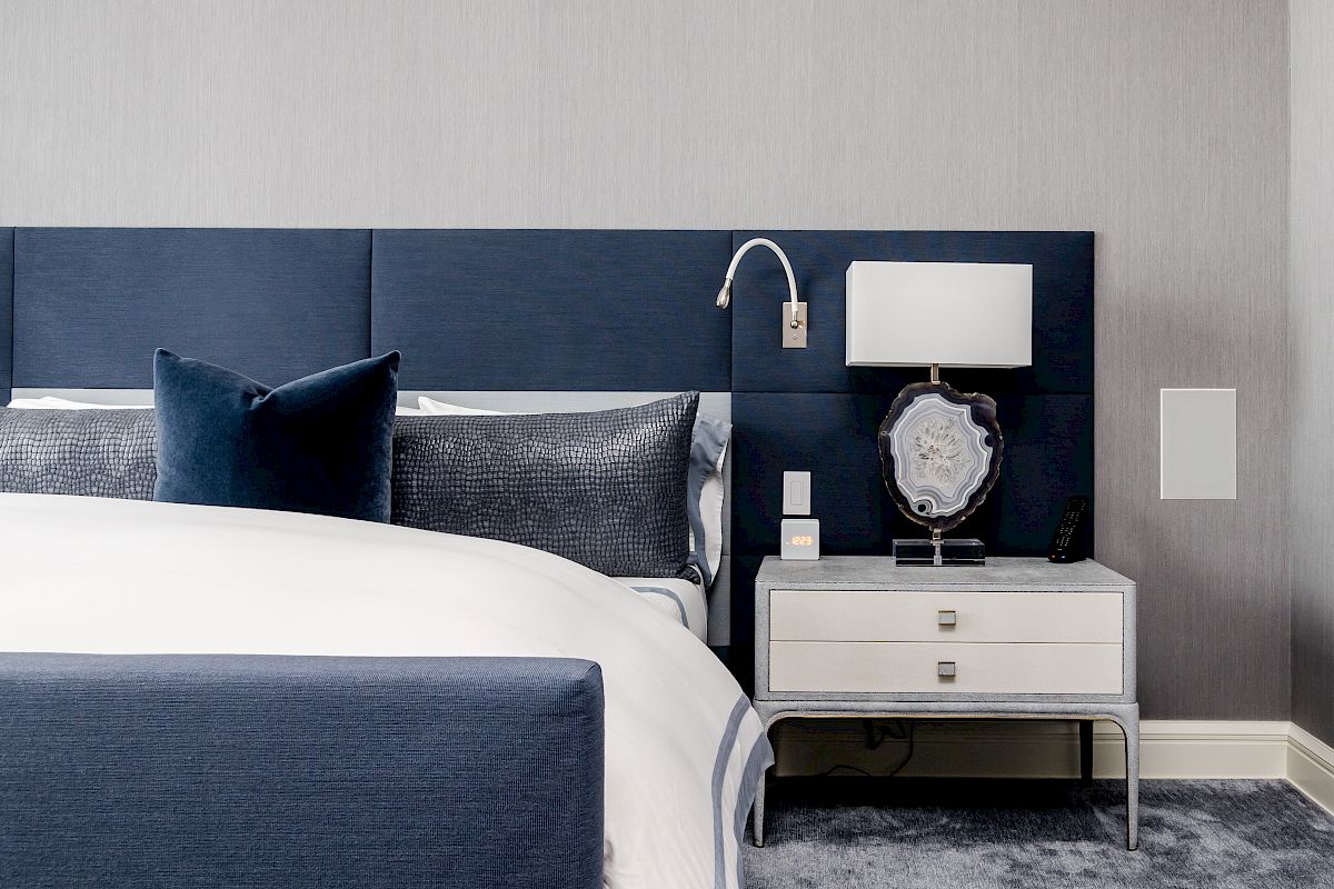 A modern bedroom with a blue upholstered bed, navy pillows, a white nightstand with a lamp and decor, on a gray carpeted floor.