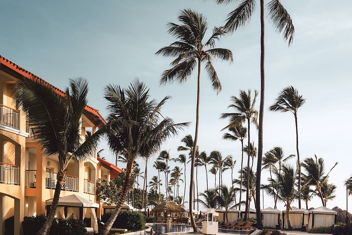 A luxurious poolside scene with palm trees, lounge chairs, and a hotel building under a clear sky.