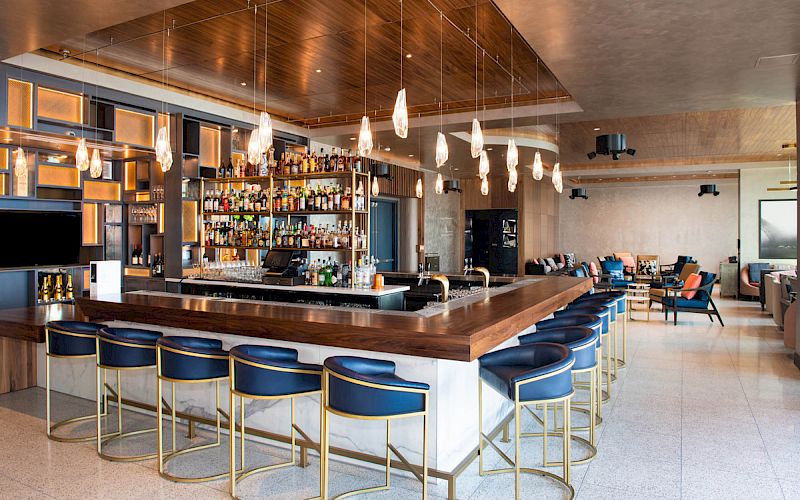 A modern bar with a sleek wooden counter, hanging lights, blue cushioned stools, and a wide variety of bottles on display in the background.