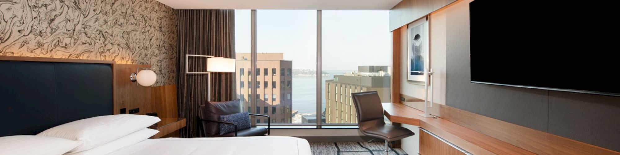 The image shows a modern hotel room with a large bed, a desk, and a flat-screen TV. The window offers a view of urban buildings.